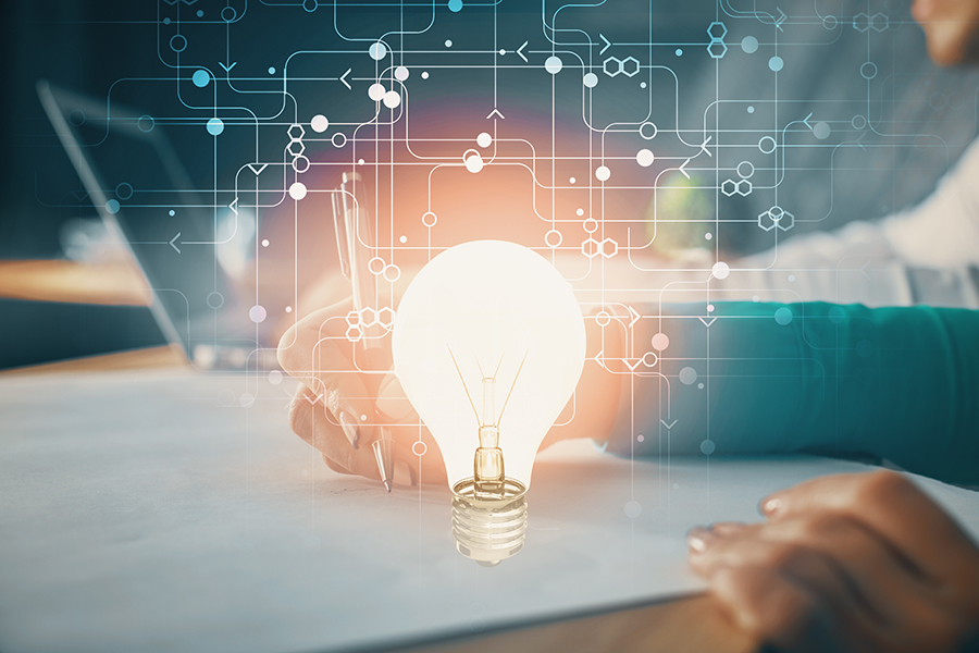 A person's hands holding and writing on a piece of white paper. There is a lit-up lightbulb where the person's pencil is.